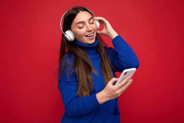 Foto von schönen fröhlich lächelnde junge Frau trägt stilvolle lässige Kleidung isoliert über Hintergrundwand halten und mit Handy tragen weiße Bluetooth-Kopfhörer Musik hören und - Foto, Bild