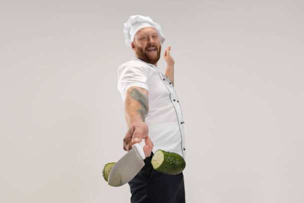 Unique cutting of vegetables. One hadsome bearded man, cook, male chef in white uniform cut cucumber isolated on white background. - 写真・画像