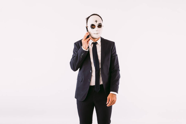 Man dressed in a suit and tie, wearing a Halloween cross on his forehead killer mask, talking on his mobile phone. Carnival and halloween celebration - Foto, Imagen