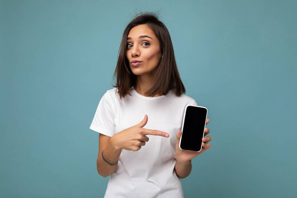 魅力的な若いブルネットの女性良い見て着て白いTシャツ立って青の背景に隔離されたコピースペースを保持スマートフォンを手に空の画面表示とモックアップのための画面表示 - 写真・画像