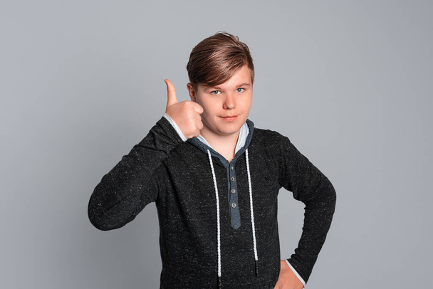Hezký teenager Gesturing Thumb Up, usměvavý a šťastný za úspěch, izolovaný na šedém pozadí, oblečený v neformálním oblečení. Koncept výrazu a emocí - Fotografie, Obrázek