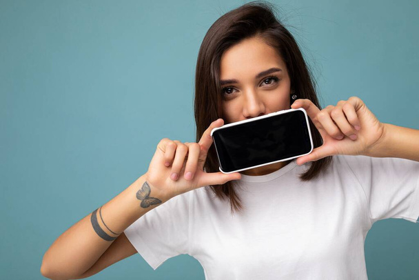 detailní fotografie docela pozitivní mladé brunetky žena dobře vypadající na sobě bílé tričko stojící izolované na modrém pozadí s kopírovat prostor drží smartphone ukazující telefon v ruce s prázdnou obrazovkou - Fotografie, Obrázek