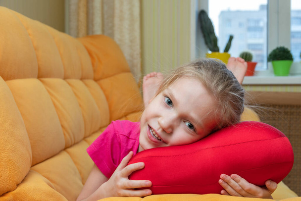 Смешная и милая блондинка, маленькая смеющаяся девочка, лежащая на красной подушке на оранжевом диване в детской комнате. Дети весёлые эмоции. Концепция счастливого ребенка. - Фото, изображение
