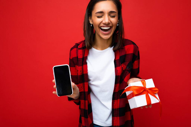 Shot of beautiful positive laughing young brunette woman isolated over red background wall wearing white casual t-shirt and red and black shirt holding white gift box with red ribbon and mobile phone - Фото, зображення