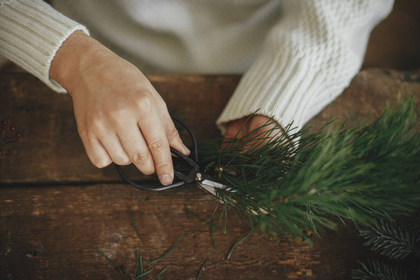Making christmas wreath. Woman in cozy sweater cutting pine branch with scissors on rustic wooden background, hands close up. Winter holiday preparations. Festive workshop, handmade decor - Φωτογραφία, εικόνα