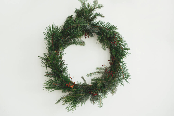Elegante corona de Navidad rústica colgando de la pared blanca en la habitación escandinava. Feliz Navidad y Felices Fiestas! Corona de Navidad creativa con ramas de pino y bayas rojas aisladas en blanco - Foto, Imagen