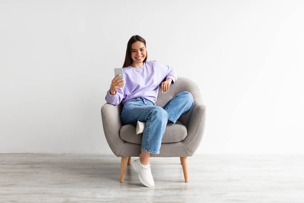 Smiling millennial woman chatting online on smartphone, sitting in comfy armchair on white studio background - Foto, immagini