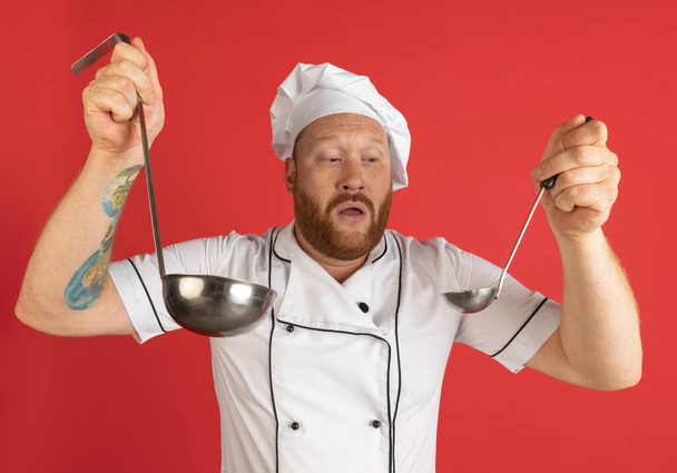 Comic portrait of hadsome bearded man, cook, male chef in white uniform having fun isolated on red studio background. Concept of job, occupation, humor. - Zdjęcie, obraz