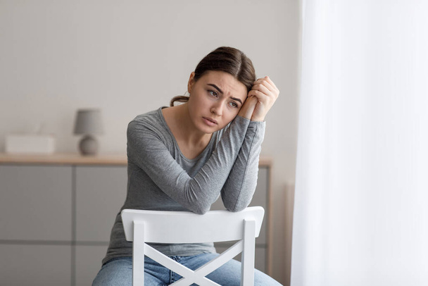 Αναστατωμένη νεαρή γυναίκα κάθεται μόνη της, κοιτάζοντας μακριά με θλιβερή έκφραση στο εσωτερικό του σπιτιού - Φωτογραφία, εικόνα