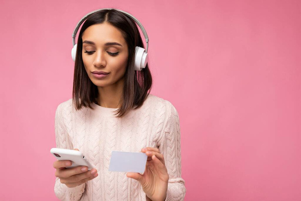Φωτογραφία από την όμορφη συμπυκνωμένη νεαρή μελαχρινή γυναίκα φορώντας ροζ casual πουλόβερ απομονωμένη πάνω από ροζ φόντο τοίχο φορώντας λευκά ακουστικά bluetooth ασύρματα και ακούγοντας μουσική και χρησιμοποιώντας κινητό - Φωτογραφία, εικόνα