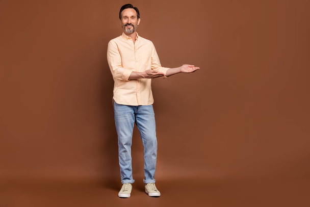 Photo of aged guy palms demonstrate empty space wear beige shirt jeans shoes isolated brown color background - Foto, immagini