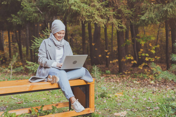 秋の公園でノートパソコンを使う若いアジア系ムスリム女性。オンライン教育、オンラインショッピング、ソーシャルネットワークの概念. - 写真・画像
