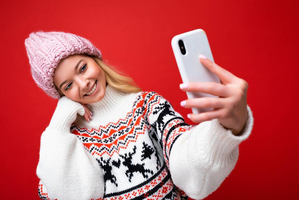 Aantrekkelijke charmante jonge glimlachende gelukkige vrouw houden en het gebruik van mobiele telefoon selfie het nemen van stijlvolle kleren geïsoleerd over muur achtergrond - Foto, afbeelding