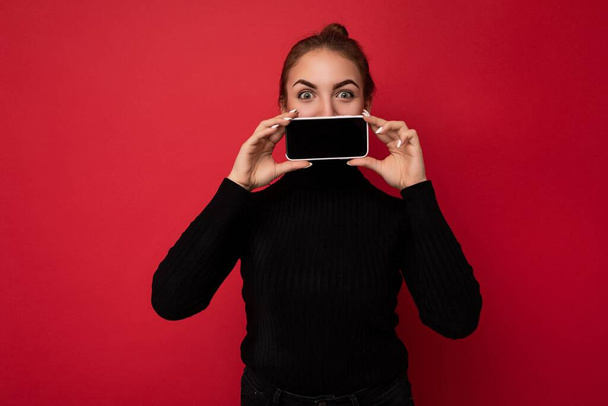 Photo of attractive positive young brunette woman wearing black sweater standing isolated over red background showing mobile phone with empty screen for mockup looking at camera - Photo, Image