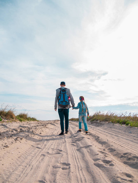 Ευτυχισμένος πατέρας και κόρη τρέχουν πηδώντας διασκεδάζοντας σε άδεια φθινοπωρινή παραλία. Ο μπαμπάς και το παιδί περπατώντας σε λευκό δρόμο άμμο με καλάμια μπλε φόντο του ουρανού. Αληθινοί άνθρωποι. Έννοια ταξιδιού ευτυχίας - Φωτογραφία, εικόνα
