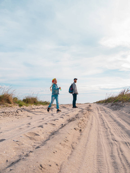 Boldog apa és lánya fut ugrás szórakozás üres őszi tengerparton. Apa és a gyerek sétálnak a fehér homokos úton nádas kék ég háttér. Igazi emberek életstílusa. Boldogság utazási koncepció - Fotó, kép