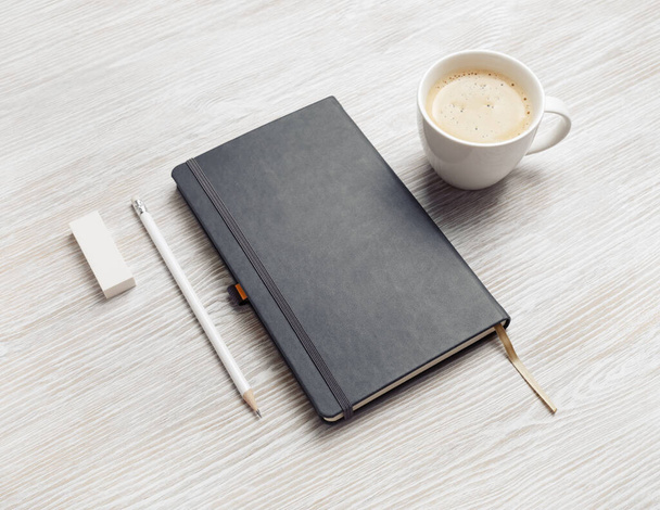 Blanco gesloten notitieboek, potlood, gum en koffiebeker op lichte houten tafelondergrond. - Foto, afbeelding
