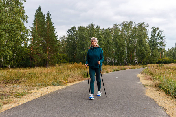 slender woman of retirement age is engaged in Nordic walking in the park. - Фото, изображение