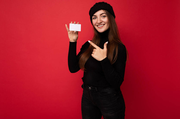 Foto van mooie jonge brunette vrouw dragen zwarte trui en hoed geïsoleerd op rode achtergrond met credit card kijken naar de camera en wijzen vinger naar plasctische kaart - Foto, afbeelding