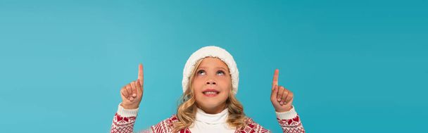 smiling child in warm hat looking up and pointing with fingers isolated on blue, banner - Foto, afbeelding
