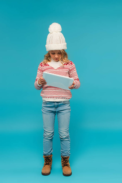 full length view of girl in jeans, knitted hat and stylish sweater using digital tablet on blue - Zdjęcie, obraz