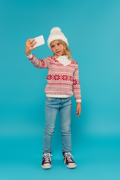 full length view of girl in warm sweater and jeans taking selfie on smartphone on blue - Foto, afbeelding
