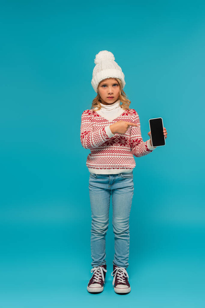 child in knitted hat, jumper and jeans pointing at smartphone with blank screen on blue - Fotografie, Obrázek