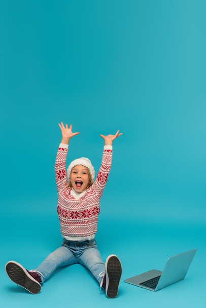 excited girl screaming and gesturing with raised hands near laptop on blue - Photo, Image