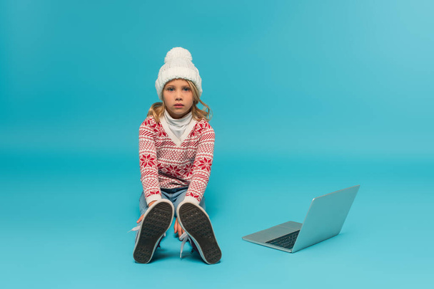 dziecko w dzianiny kapelusz i sweter z ornament siedzi w pobliżu laptopa na niebiesko - Zdjęcie, obraz