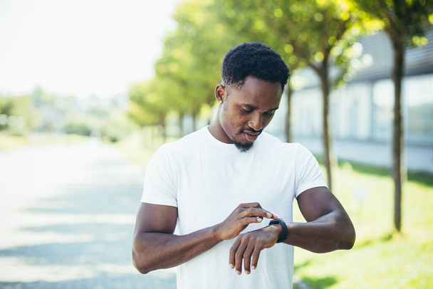 Young african american man, on a morning jog in a white t-shirt in the park, switches sports program in smart watch fitness bracelet engaged in fitness active lifestyle - Φωτογραφία, εικόνα