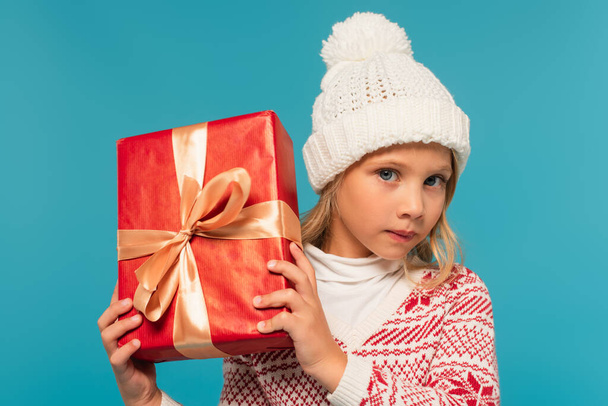 kid in knitted hat and sweater looking at camera while holding red gift box isolated on blue - Photo, Image