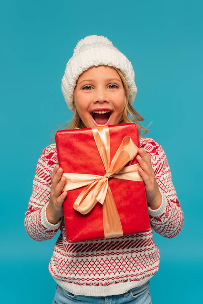 excited girl in warm hat looking at camera while holding red gift box isolated on blue - Foto, Bild