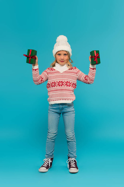 vista completa de niño en suéter y jeans mostrando cajas de regalo verdes en azul - Foto, Imagen