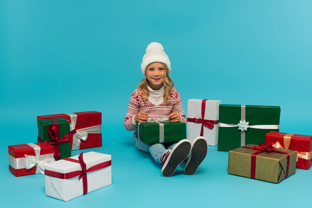 gelukkig meisje in warm hoed en trui kijken naar camera terwijl zitten in de buurt cadeautjes op blauw - Foto, afbeelding
