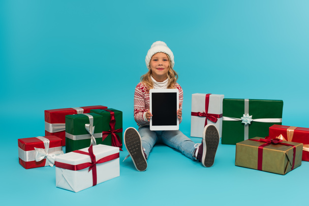 szczęśliwy dzieciak w swetrze i kapelusz siedzi z tabletem cyfrowym w pobliżu wielobarwnych pudełek upominkowych na niebiesko - Zdjęcie, obraz