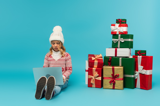 dítě v pleteném klobouku a svetru sedí u hromady dárků a pomocí notebooku na modré - Fotografie, Obrázek