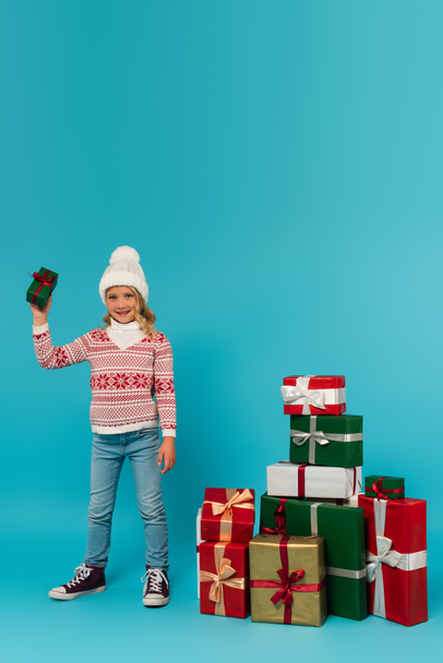 child in warm hat, sweater and jeans holding present near stack of gift boxes on blue - Φωτογραφία, εικόνα