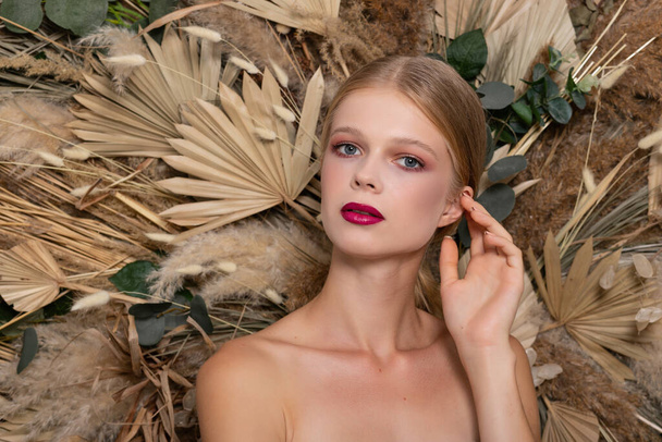 looks straight and hand fozle face Blonde girl with burgundy lips against a background of spring dry field flowers. Beauty face care concept. - 写真・画像