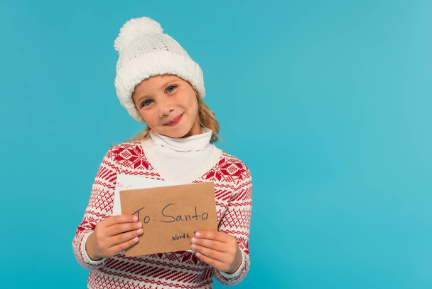 joyful child in knitted hat and sweater showing letter to santa isolated on blue - Fotografie, Obrázek