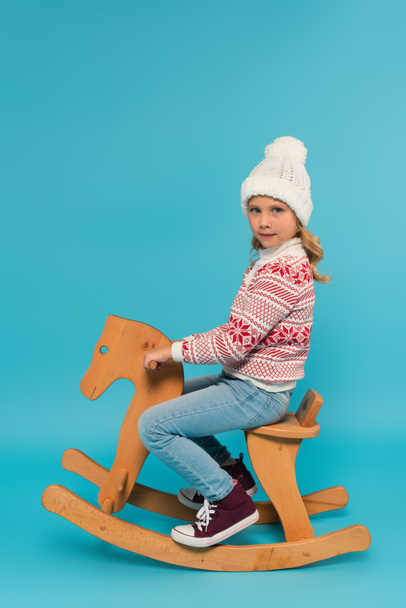 girl in warm hat and sweater riding rocking horse while looking at camera on blue - Фото, изображение