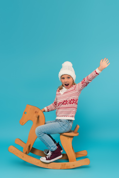 excited child in knitted hat and jumper shouting and waving hand on rocking horse on blue - 写真・画像