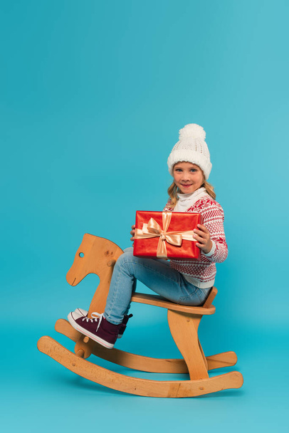 joyful girl in knitted hat and sweater holding present while riding rocking horse on blue - Photo, Image
