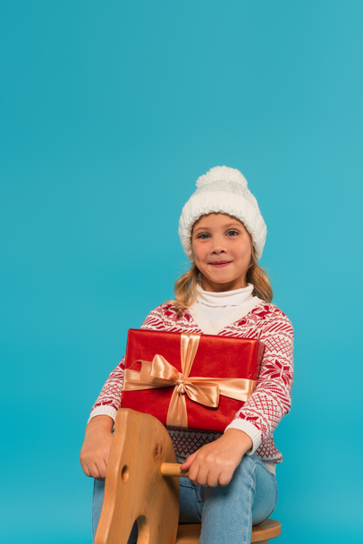 pleased girl in knitted hat looking at camera while riding rocking horse with gift box isolated on blue - Fotografie, Obrázek