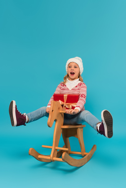 cheerful girl in warm hat shouting while riding rocking horse with gift box on blue - Photo, Image