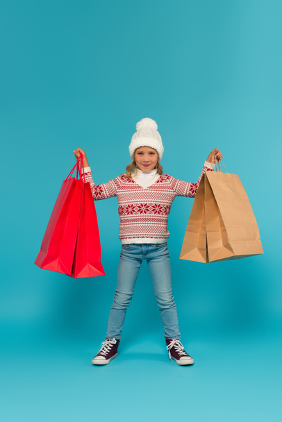 child in warm sweater, jeans and sneakers standing with shopping bags on blue - Фото, зображення