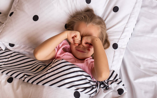 Маленькая блондинка в пижаме просыпается в постели и массирует глаза руками. - Фото, изображение