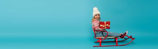 girl in warm hat and sweater sitting in sled with red gift box on blue, banner - Valokuva, kuva