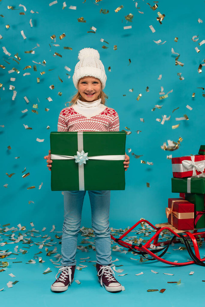 joyful kid in warm hat holding big gift box near confetti and sled with presents on blue - Photo, image