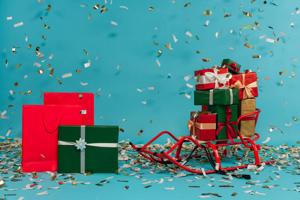 sled with presents near shopping bags and gift boxes under falling confetti isolated on blue - Foto, immagini