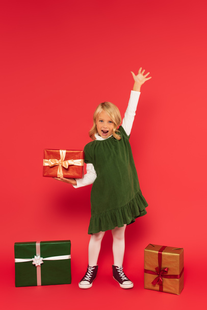 astonished girl holding present and shouting while standing with raised hand near gift boxes on red - Foto, Imagem
